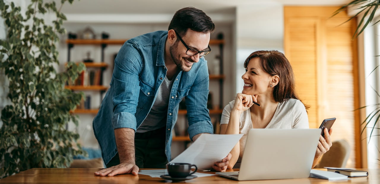 A smiling couple reviews a document; there is a laptop before them on a table